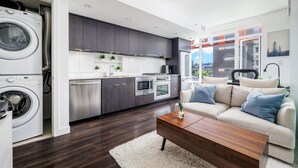 A modern kitchen with built-in appliances, a dining area with a sofa and coffee table, and a large window offering a city view. - FIFA Stay 1BR| Steps to Seawall | Free Parking+AC (Vancouver)