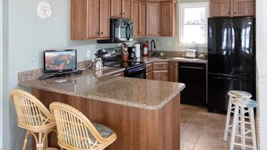 Una cocina con armarios de madera, encimera de granito, un refrigerador negro y dos taburetes de mimbre.