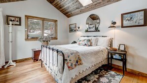 A bedroom with a wooden ceiling, a bed with a quilt, a bedside table, a mirror, and a window with a view of trees. - Crooked Pine Cottage- Close To RMNP Entrance (Estes Park)