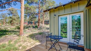 A wooden deck with a table and chairs, a glass door with a view of trees, and a building with a green exterior. - Crooked Pine Cottage- Close To RMNP Entrance (Estes Park)