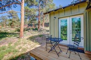 A wooden deck with a table and chairs, a glass door with a view of trees, and a building with a green exterior.