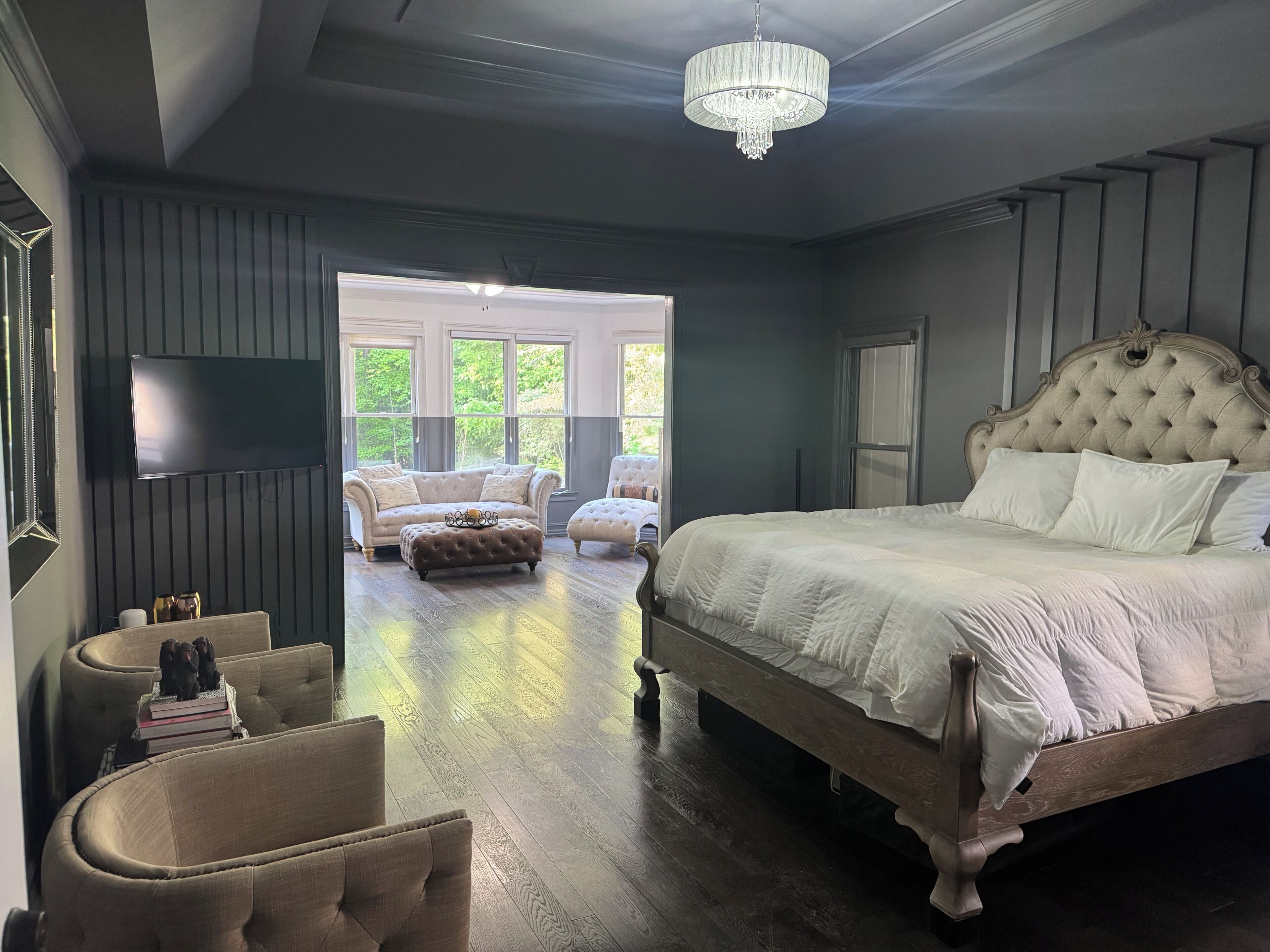 A bedroom with a large bed, a television, a chandelier, and a view of the outdoors.