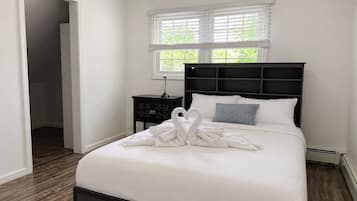 A neatly made bed with white linens and a heart-shaped towel arrangement. There is a window with blinds, a bedside table with a lamp, and a dark headboard.