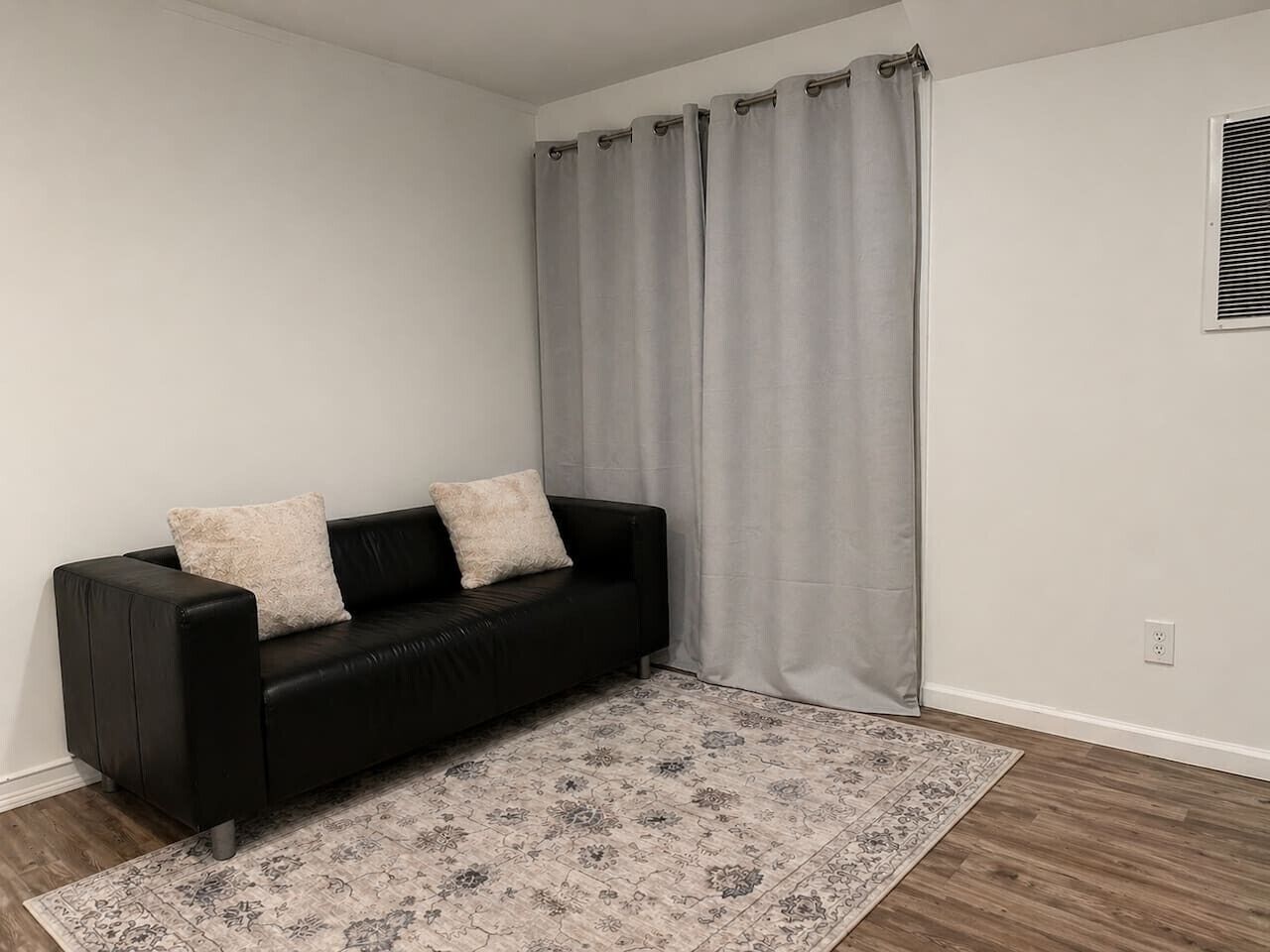 Un sofá de cuero negro con dos cojines, un tapete estampado y una cortina gris en una habitación con paredes blancas y suelo de madera.