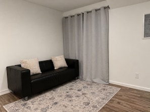 A black leather sofa with two cushions, a patterned rug, and a grey curtain in a room with white walls and a wooden floor. - North Brunswick 2BR | Easy Access to NYC & Rutgers (North Brunswick Township)
