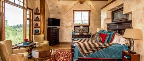 A spacious bedroom with a large bed, a sitting area with a chair and ottoman, a television, and a fireplace.