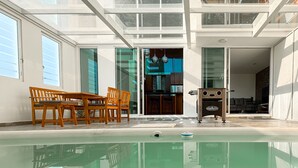 Un espace piscine intérieur moderne, doté de murs en verre, d’une table et de chaises en bois, et d’une cuisine visible à travers une porte ouverte.