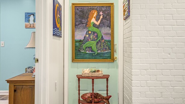 Un pasillo con pared color turquesa, un cuadro enmarcado de una sirena, una mesa de madera con una cesta y una escultura de pez.