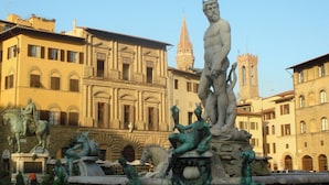 A classical fountain with statues in front of a building with arched windows and a tower.