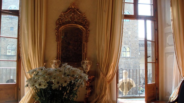 A room with a large mirror, a vase of flowers, and a view of a building through large windows.