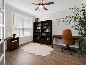 Ein Zimmer mit Deckenventilator, Bücherregal, Schreibtisch, Stuhl, Fenster mit Jalousien und einer Wand mit motivierendem Zitat.