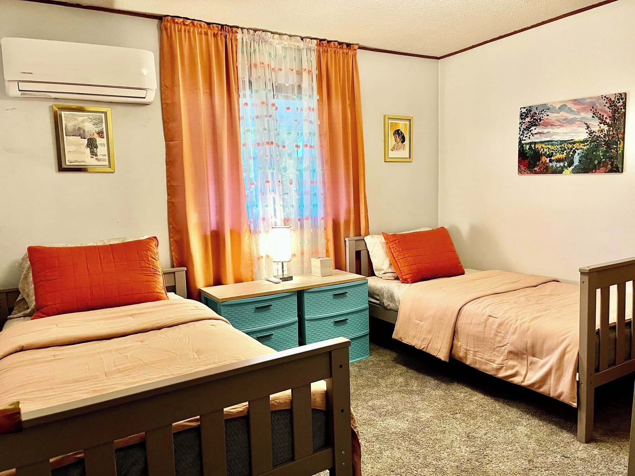 Un dormitorio con dos camas, una mesita de noche, un aparador, un aire acondicionado y dos cuadros en la pared.