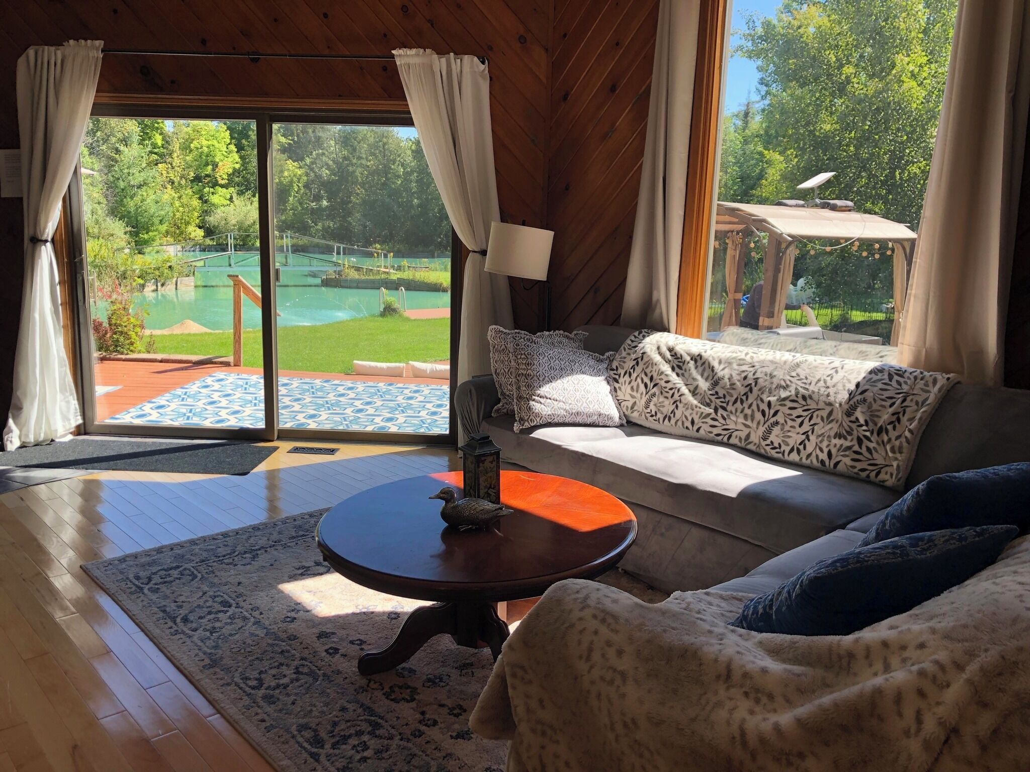 Une pièce avec un plafond en bois, un canapé agrémenté de coussins à motifs, une table ronde avec une lampe, et une vue sur une piscine à travers de grandes portes vitrées.