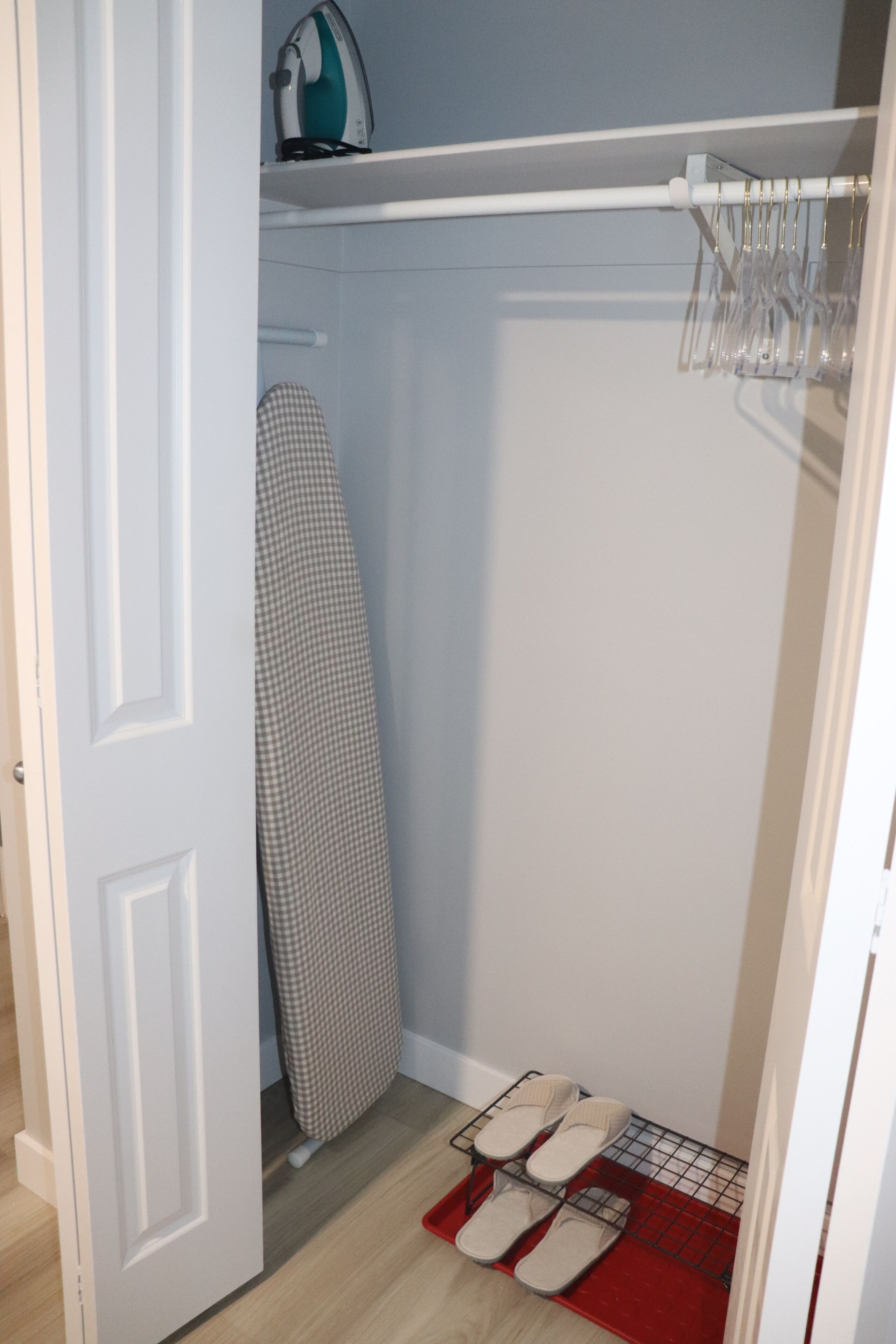A walk-in closet with an ironing board, a hanging rack, and a pair of slippers on a red mat.