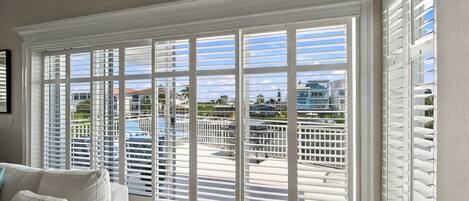 A room with a large window with white blinds, a sofa, and a view of a pool and buildings.