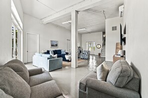 Amplia sala de estar con un sofá grande, una mesa de centro y un sillón estampado. Se ve una escalera que sube a otro nivel, y una puerta a la izquierda.