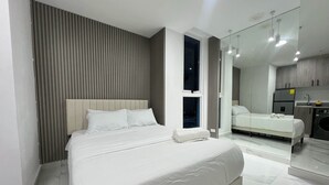 Una habitación de hotel moderna con una cama grande, una kitchenette pequeña y un baño visible a través de un separador de vidrio.