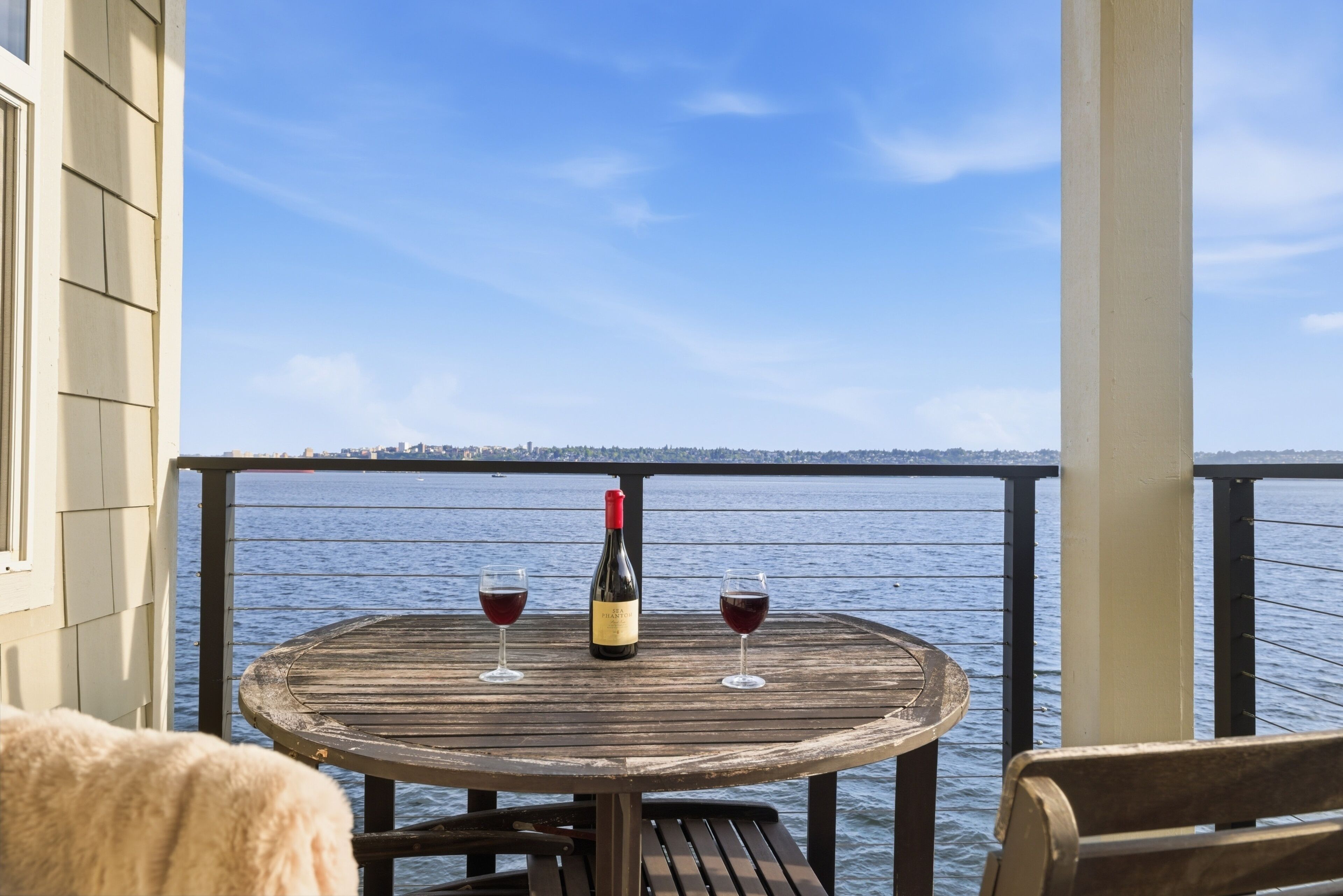 A wooden table with two wine glasses and a bottle on a balcony overlooking a body of water.