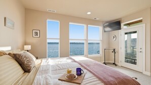 A bedroom with a bed, a bedside table with a lamp, a window with a view of the water, and a door with a TV mounted above it. - Puget Sound waterfront · Rainier · fireplace (Tacoma)