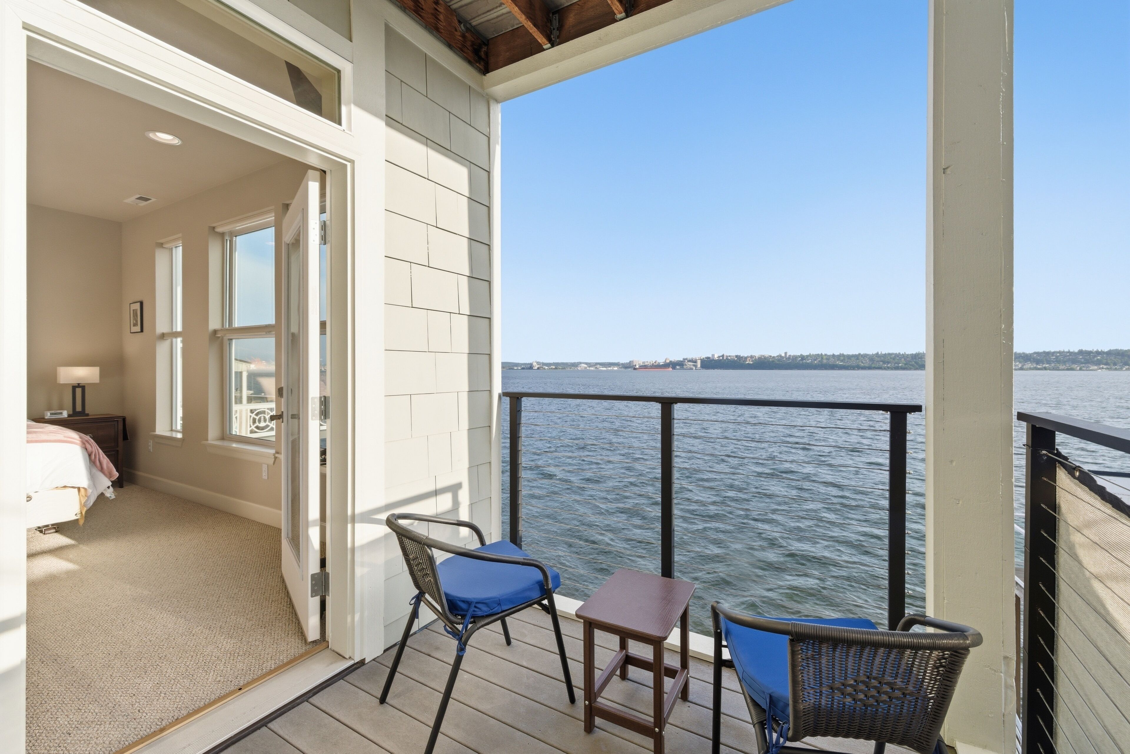 A balcony with a view of the water, a chair, a table, and a bedroom with a bed and nightstand.