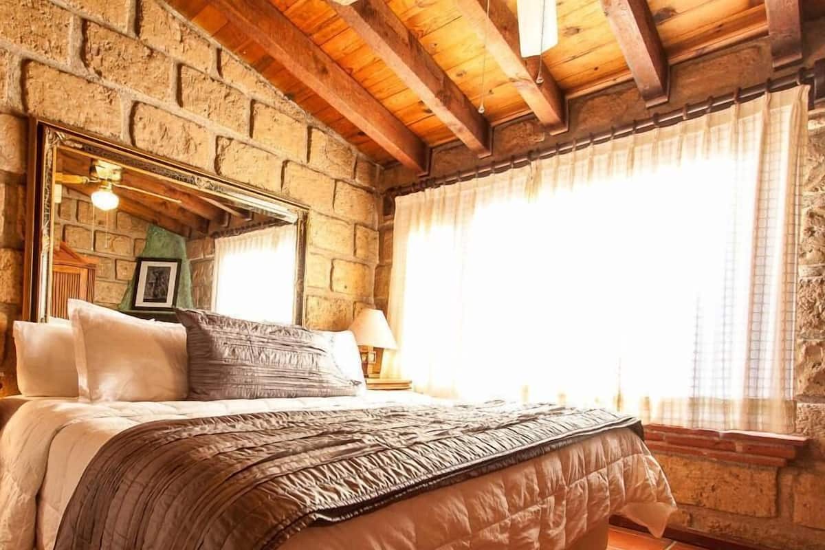 A bedroom with a stone wall, wooden beams, a bed with pillows, a lamp, and a window with curtains.
