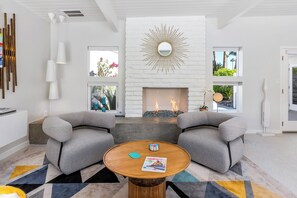 Ein modernes Wohnzimmer mit Kamin, zwei grauen Sesseln, einem runden Couchtisch und einem Teppich mit geometrischem Muster.
