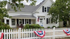 A white house with a porch, a white picket fence, and an American flag. - A quintessential Cape Cod family home in Dennis Port (Dennis)