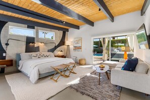Ein modernes Schlafzimmer mit einem großen Bett, Nachttischen, einem kleinen Tisch und einem Sofa. Ein Fenster bietet Blick auf Grünflächen und einen Pool.