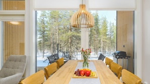 Ein Essbereich mit einem Holztisch, Stühlen, einer Vase mit Blumen und Blick auf einen Wald durch große Fenster.