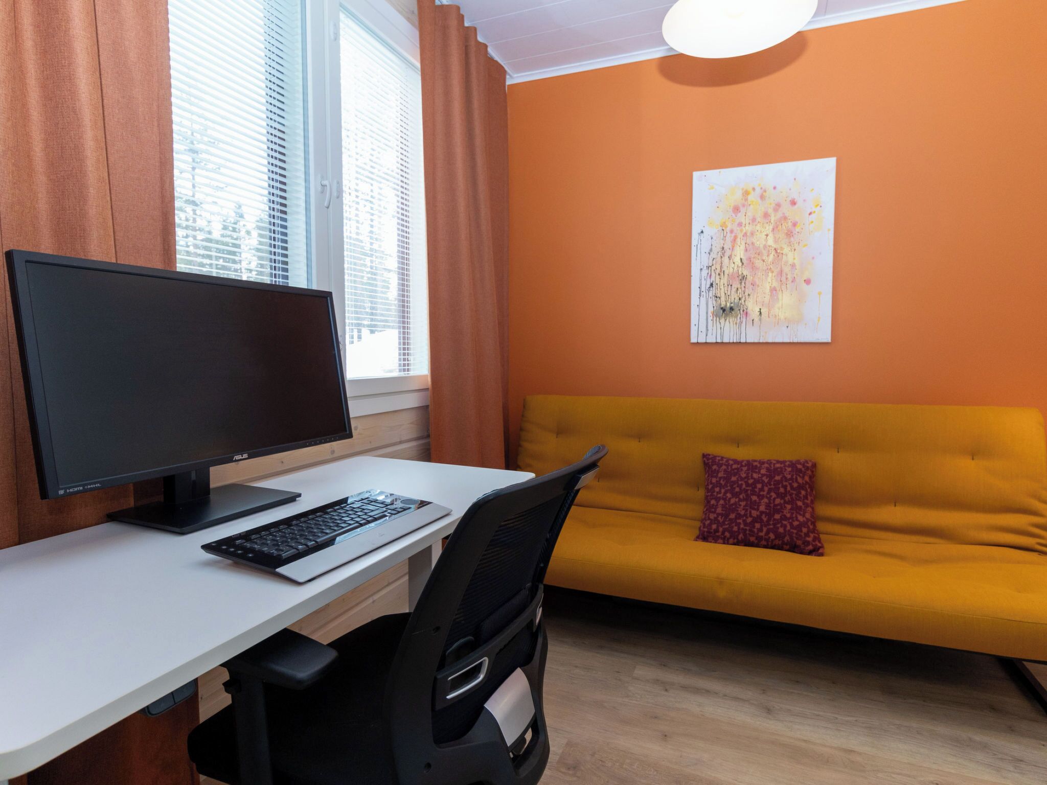 A modern room with a white desk, black monitor, and keyboard, a yellow sofa with a red pillow, and a painting on the orange wall.