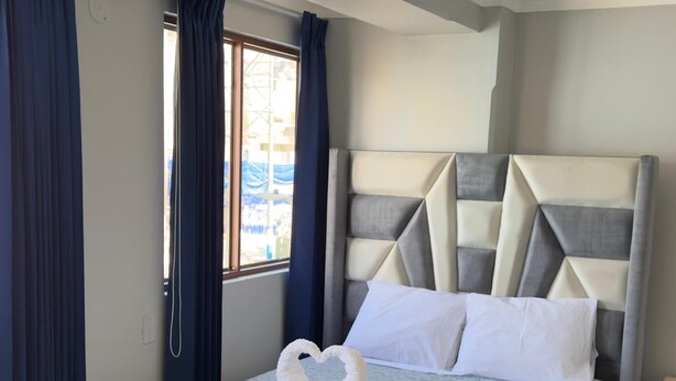 Un lit bien fait, recouvert d’une housse matelassée grise, avec une tête de lit ornée de motifs géométriques et une disposition de serviettes en forme de cœur.