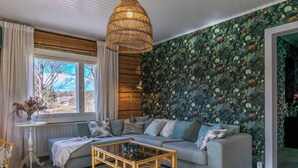 A living room with a sofa, a coffee table, a hanging light fixture, a window with curtains, and a wall with a leafy pattern. - Virtaan tainio by Interhome (Sysmä)