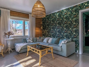 Een woonkamer met een bank, een salontafel, een hanglamp, een raam met gordijnen en een muur met een bladerenpatroon.