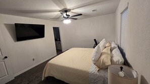 Ein Schlafzimmer mit Bett, Deckenventilator, an der Wand montiertem Fernseher und einem Fenster mit Jalousien.