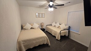 A small, well-lit bedroom with two single beds, a ceiling fan, and a window with blinds.
