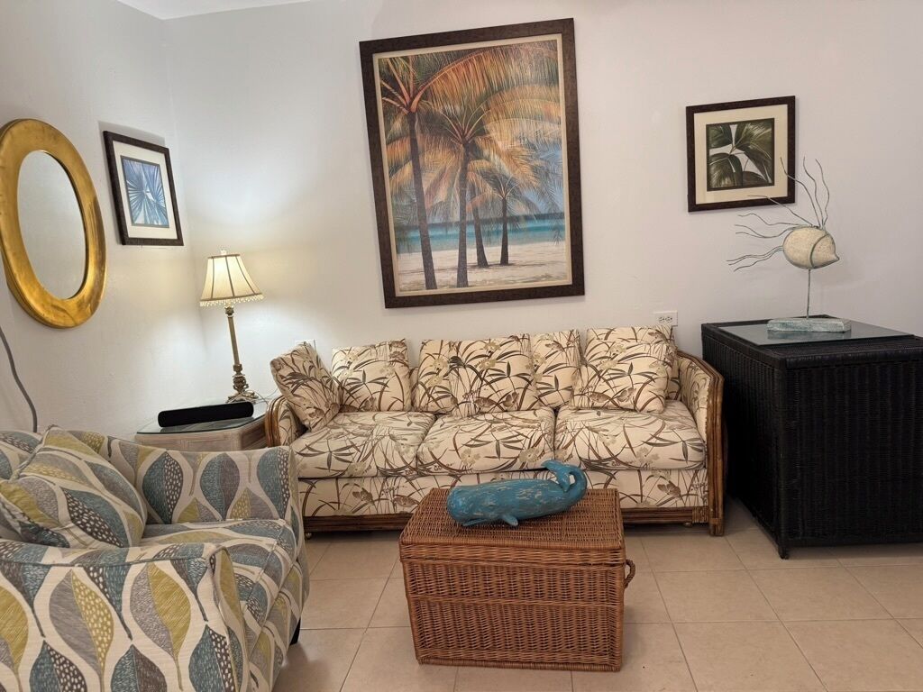 Een woonkamer met een gestreept sofa, een rieten bijzettafeltje en wanddecoratie, waaronder een strandscène en een plantbeeldhouwwerk.