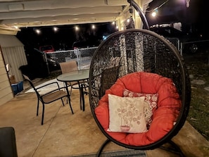 Un espace patio comprenant une chaise ronde et rembourrée ainsi qu’une petite table, le tout entouré d’une clôture et illuminé par des guirlandes lumineuses.