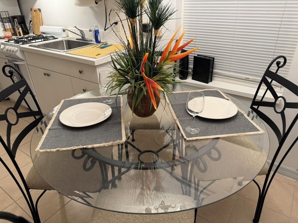 A glass-top dining table with two plates, surrounded by black metal chairs, in a kitchen with a white countertop and a potted plant.