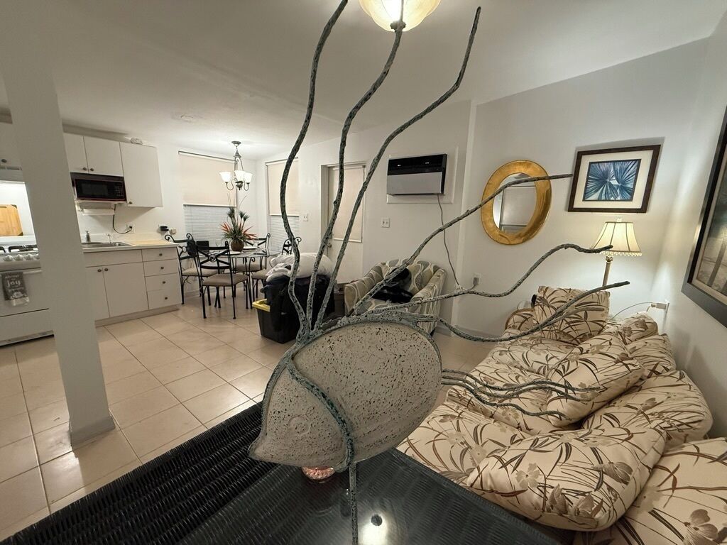Un salon moderne agrémenté d’une pièce maîtresse sculpturale originale, un coin repas illuminé par un lustre, et une cuisine dotée de meubles blancs.