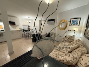 Un salon moderne agrémenté d’une pièce maîtresse sculpturale originale, un coin repas illuminé par un lustre, et une cuisine dotée de meubles blancs.