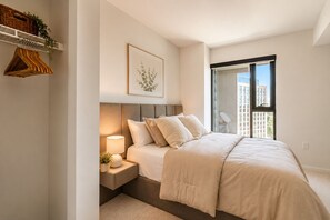 A bedroom with a bed, bedside table, lamp, and a view of buildings through a window. - Modern Luxury Full 1Bedroom Apartment. Pool, Gym & Mall, Metro, & Restaurants (Tysons)
