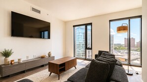 Un salon moderne doté d’un grand téléviseur à écran plat, d’une console en bois, d’un canapé gris et de grandes fenêtres offrant une vue sur la ville.