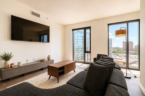 Ein modernes Wohnzimmer mit einem großen Flachbildfernseher, einem Holztischelement, einem grauen Sofa und großen Fenstern mit Blick auf die Stadt.