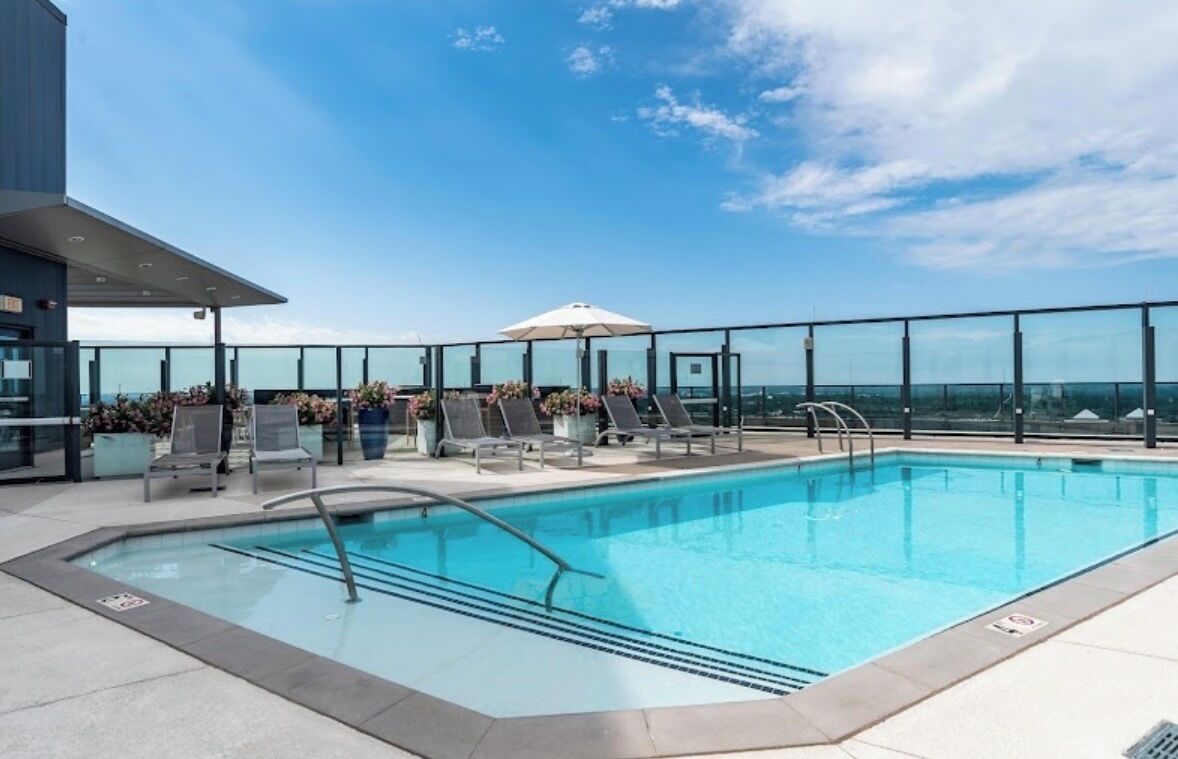 A rooftop pool with lounge chairs and a glass railing, overlooking a clear sky.