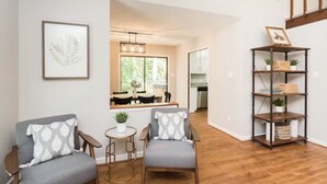 Una habitación con dos sillones grises, una pequeña mesa redonda con una planta, suelo de madera, un espejo, una estantería y una ventana con luz natural.
