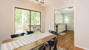 Un comedor con una mesa de madera, sillas y un jarrón de flores. Amplias ventanas y vistas al exterior con vegetación.