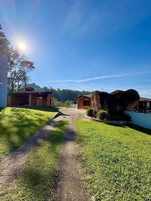 Uma trilha leva a um edifício com um grande afloramento rochoso, rodeado por gramado bem cuidado e céu claro.