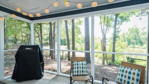 Un espace extérieur aménagé avec une zone de repos couverte, un barbecue, deux chaises dotées de coussins à motifs et une vue sur des arbres et une étendue d’eau.