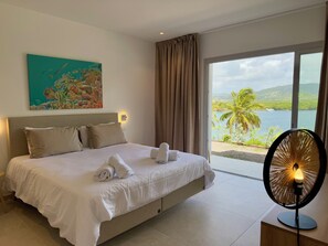 Ein Schlafzimmer mit Bett, einem Gemälde und Blick auf das Meer, sowie einem Ventilator.