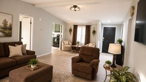 A spacious living room with a sofa, armchair, and ottoman, a dining area, and a kitchen in the background.